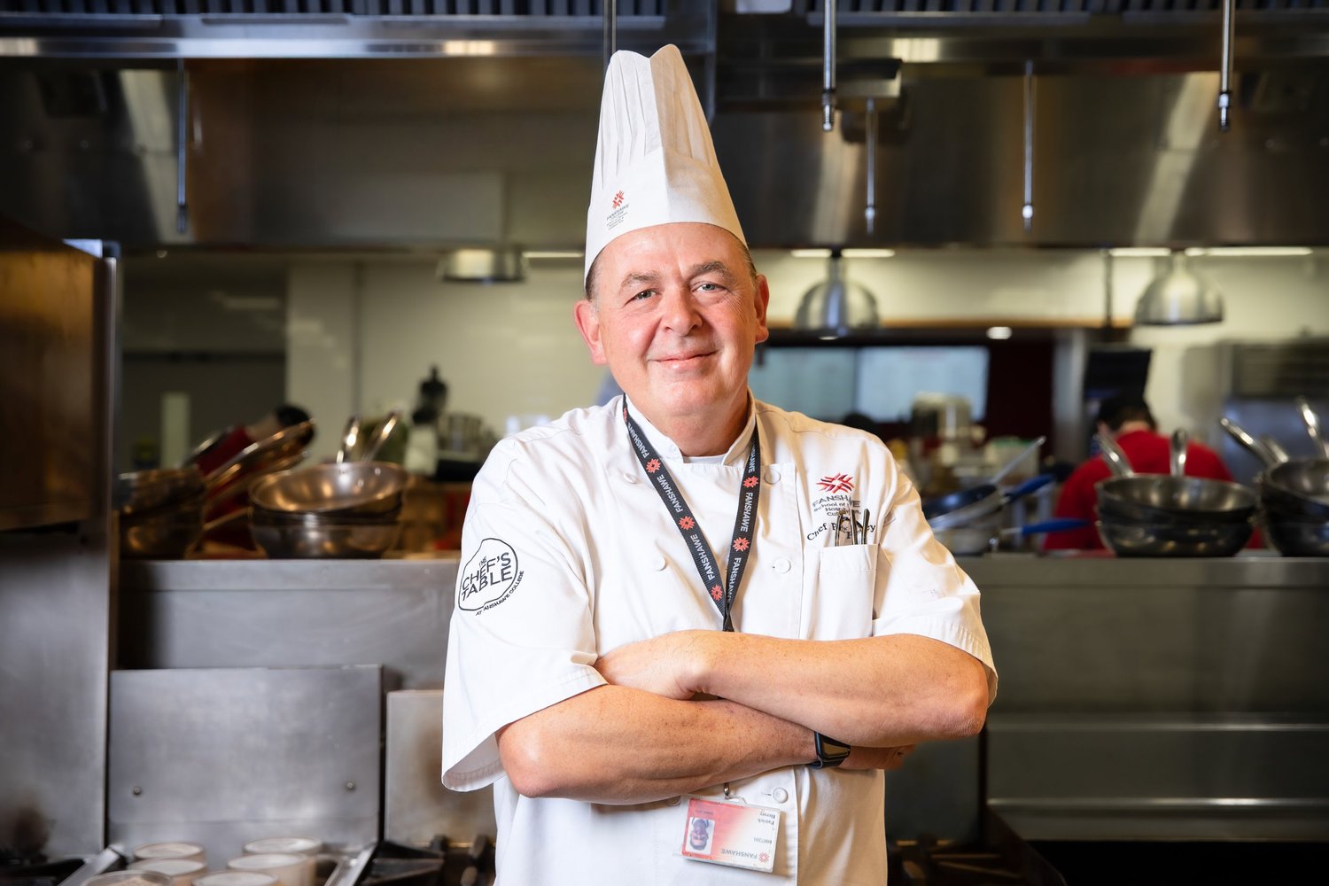 A chef in a white uniform and tall hat, standing with folded arms in a professional kitchen.